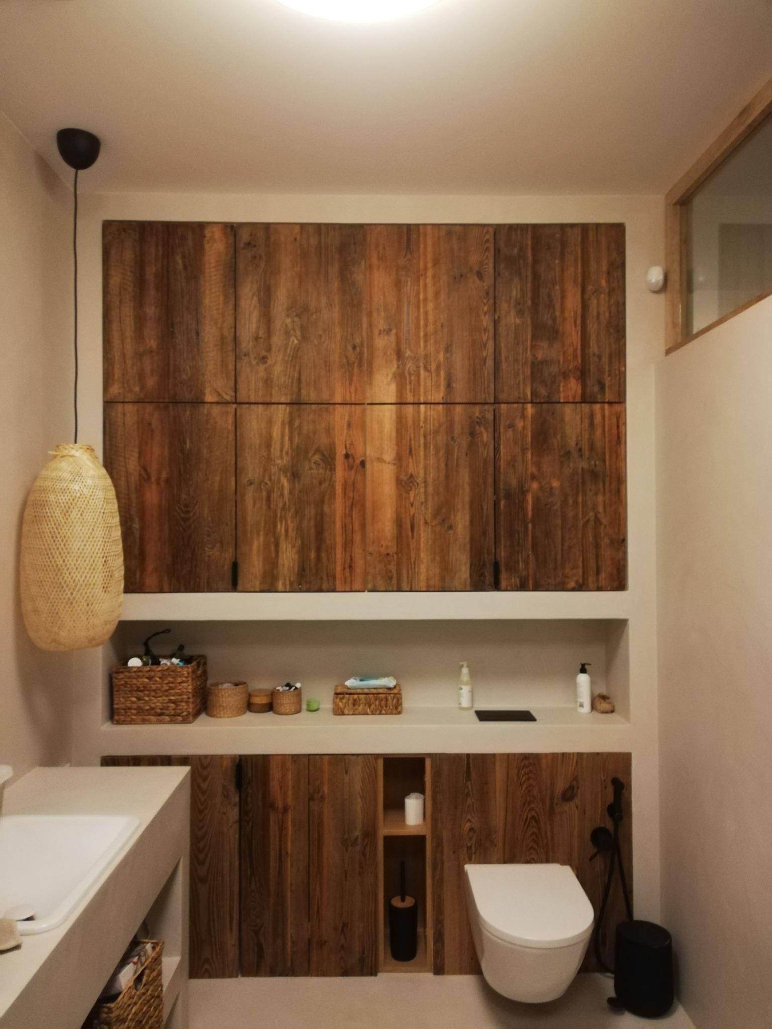 Jasne wnętrze łazienki z drewnianymi szafkami o rustykalnym wykończeniu, wisząca lampa z plecionki i umywalka z blatem z betonu architektonicznego.