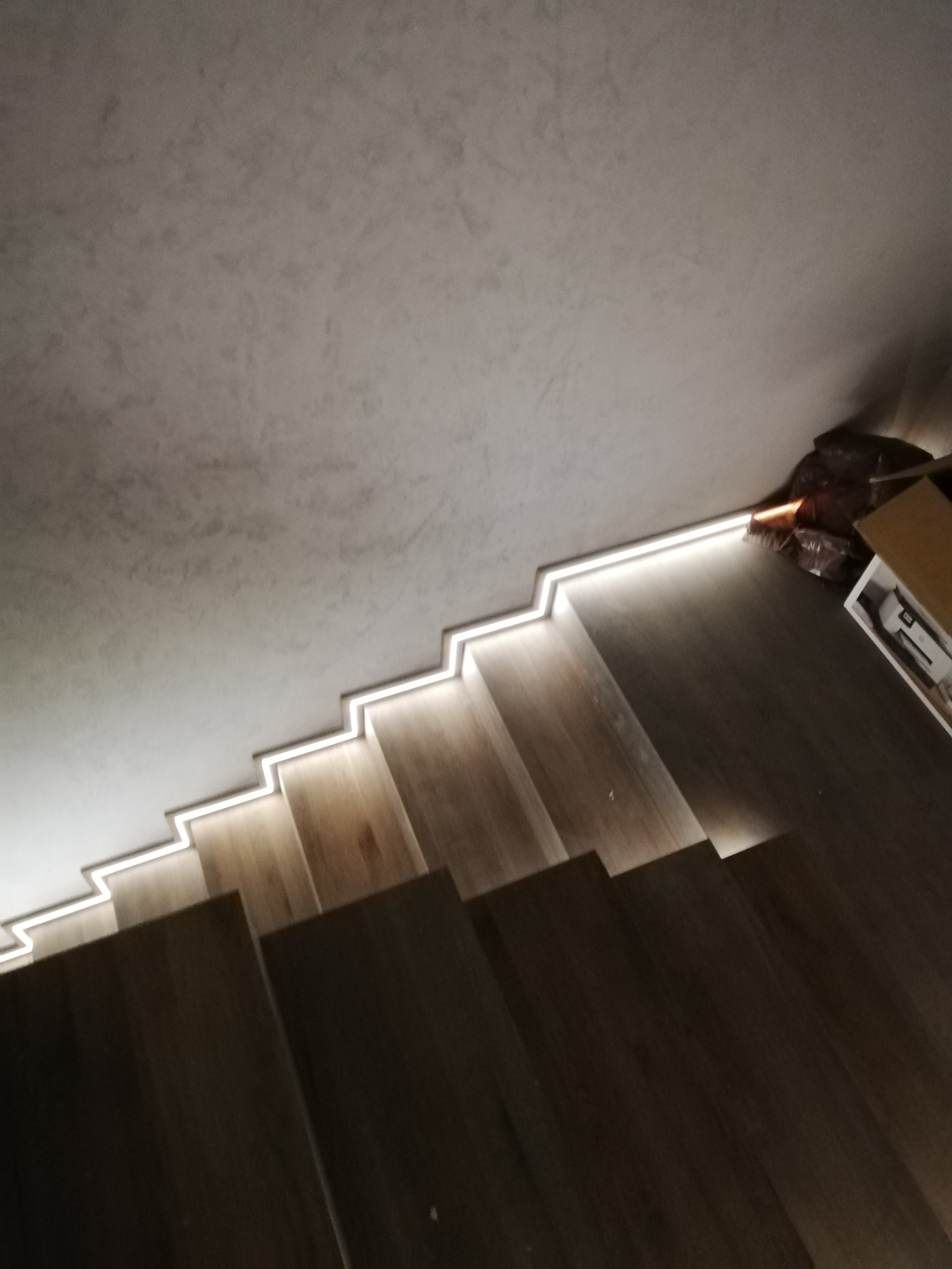 Nowoczesne schody drewniane z podświetleniem LED wzdłuż krawędzi stopni, widok z góry pod kątem, surowy betonowy sufit, częściowo widoczne materiały budowlane w tle.