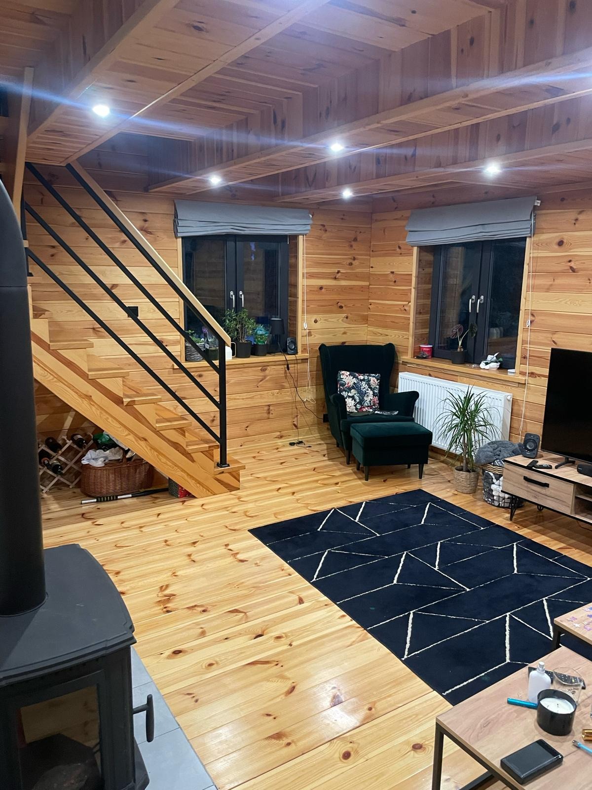 Wnętrze domu z drewnianą boazerią, schodami z czarną balustradą i czarnym dywanem w geometryczne wzory. Widoczny piec wolnostojący, fotel z podnóżkiem oraz okna z szarymi roletami rzymskimi.