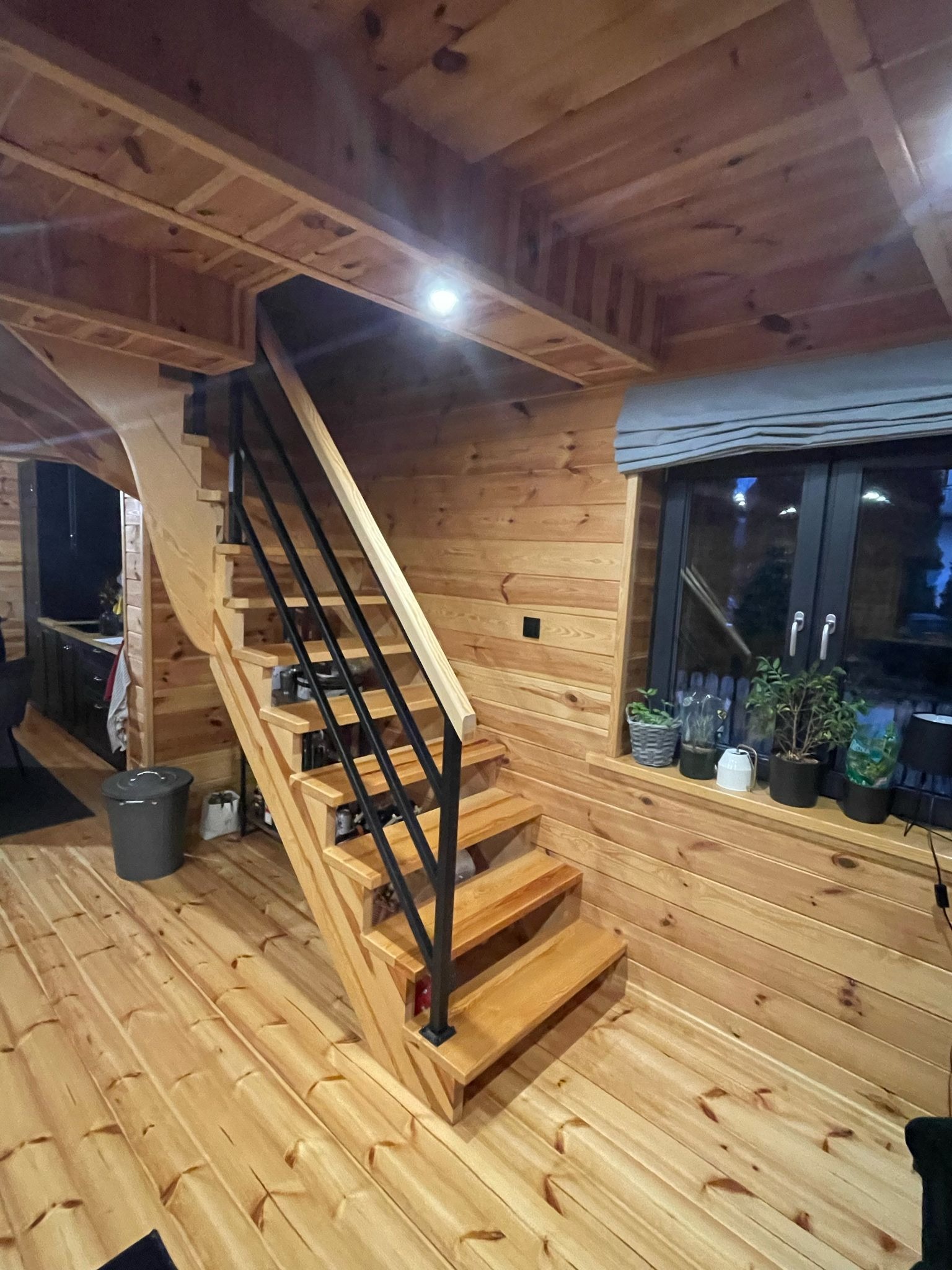 Wnętrze domu z drewnianą boazerią, schody z czarną metalową balustradą, okno z roślinami w doniczkach na parapecie.