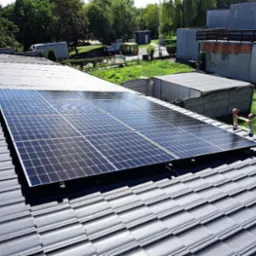 Cztery panele słoneczne na szarym dachu z dachówki ceramicznej, widok z góry, w tle budynki gospodarcze i zieleń.