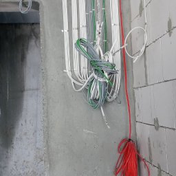 Elektro-Pal - Pionowe wiązki przewodów elektrycznych w kolorach białym, szarym, zielonym i czerwonym, przymocowane do surowej betonowej ściany i ściany z białych bloczków, widoczne kable zwisające luźno.