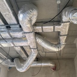 Elektro-Pal - Widok sufitu z widocznymi rurami wentylacyjnymi owiniętymi srebrną folią, przewodami elektrycznymi i czujnikami dymu, w tle stos paczek wełny mineralnej Rockwool.