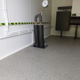 Elektro-Pal - W kotłowni w Dzierzgoniu widoczny fragment instalacji z dwoma czarnymi rurami połączonymi u góry kolankiem z manometrem, na ścianie białe rury i żółto-zielony pas ostrzegawczy, w tle piec.