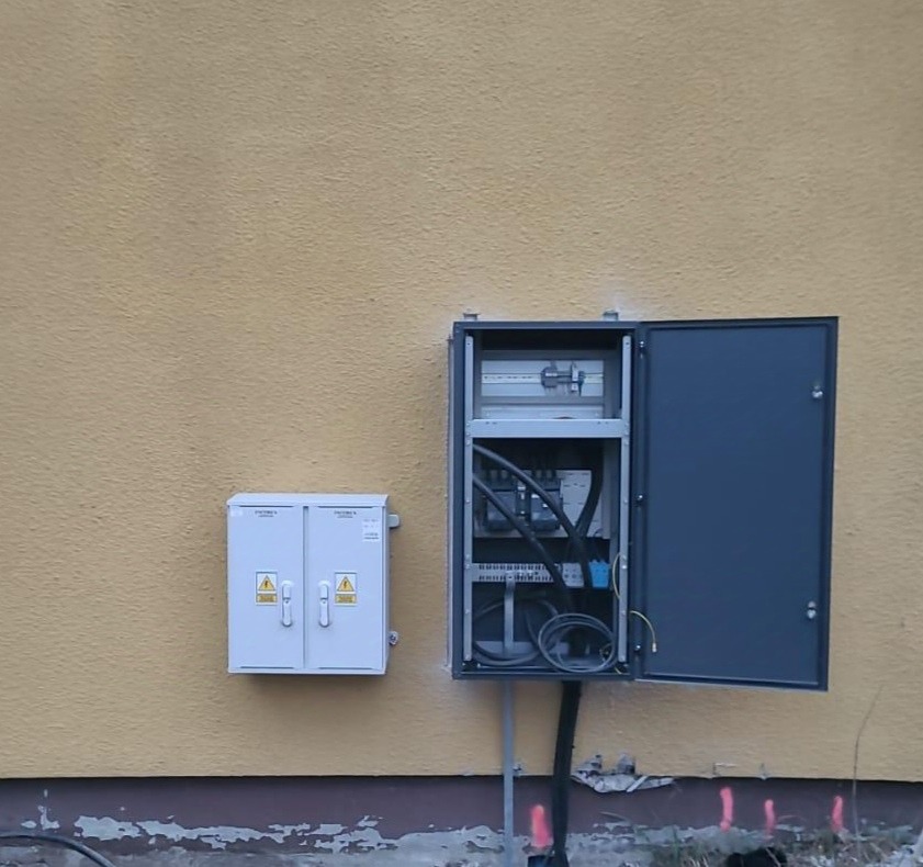 Dwie skrzynki elektryczne zamontowane na żółtej ścianie budynku; większa skrzynka z otwartymi drzwiami eksponuje wewnętrzne okablowanie i zabezpieczenia.