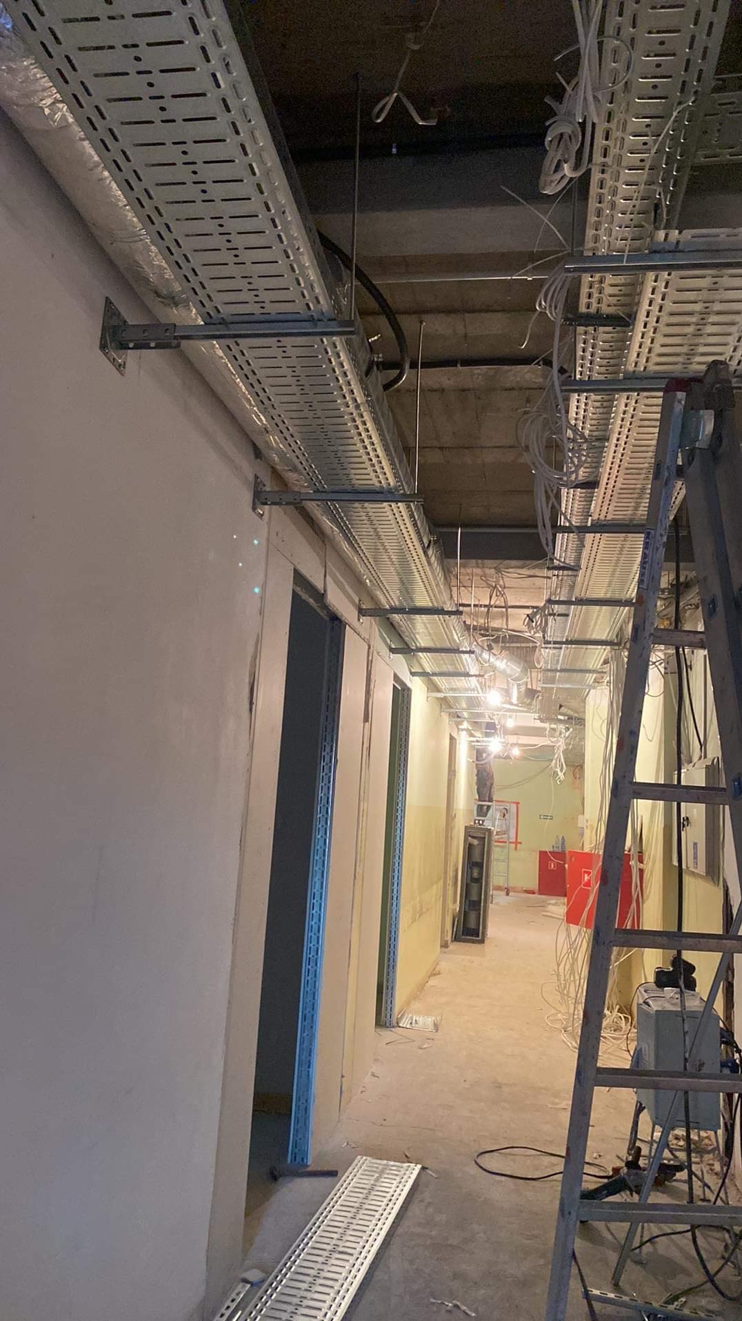 Korytarz w trakcie montażu tras kablowych pod sufitem, widoczna drabina monterska i narzędzia, surowy stan wykończenia ścian.