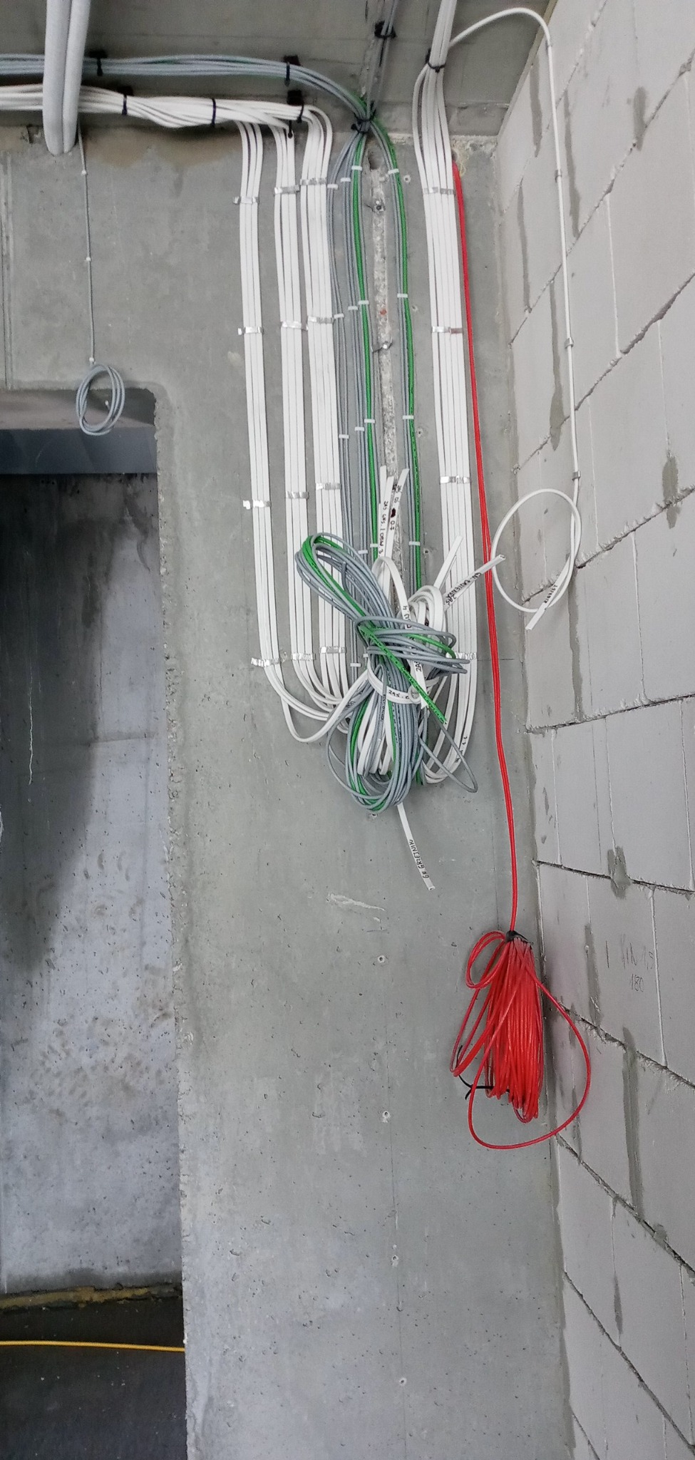 Pionowe wiązki przewodów elektrycznych w kolorach białym, szarym, zielonym i czerwonym, przymocowane do surowej betonowej ściany i ściany z białych bloczków, widoczne kable zwisające luźno.