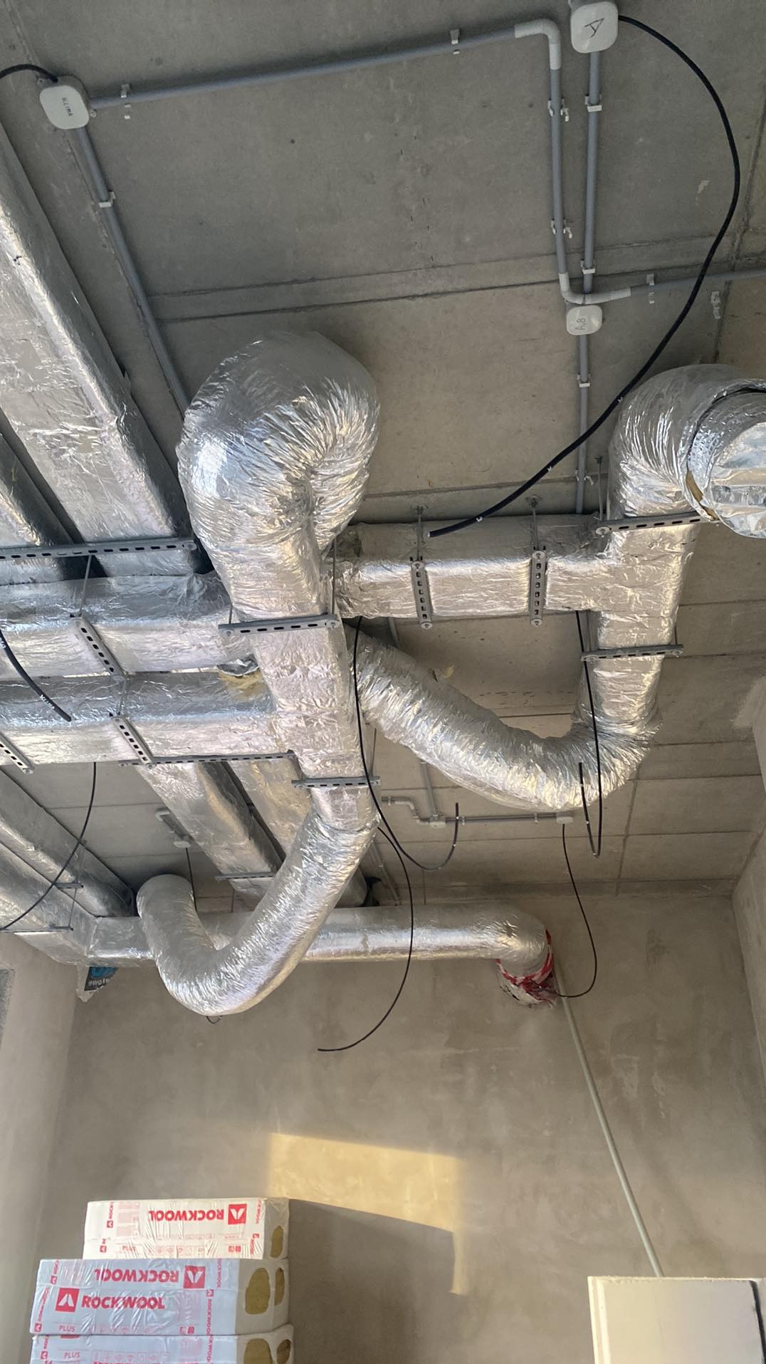 Widok sufitu z widocznymi rurami wentylacyjnymi owiniętymi srebrną folią, przewodami elektrycznymi i czujnikami dymu, w tle stos paczek wełny mineralnej Rockwool.