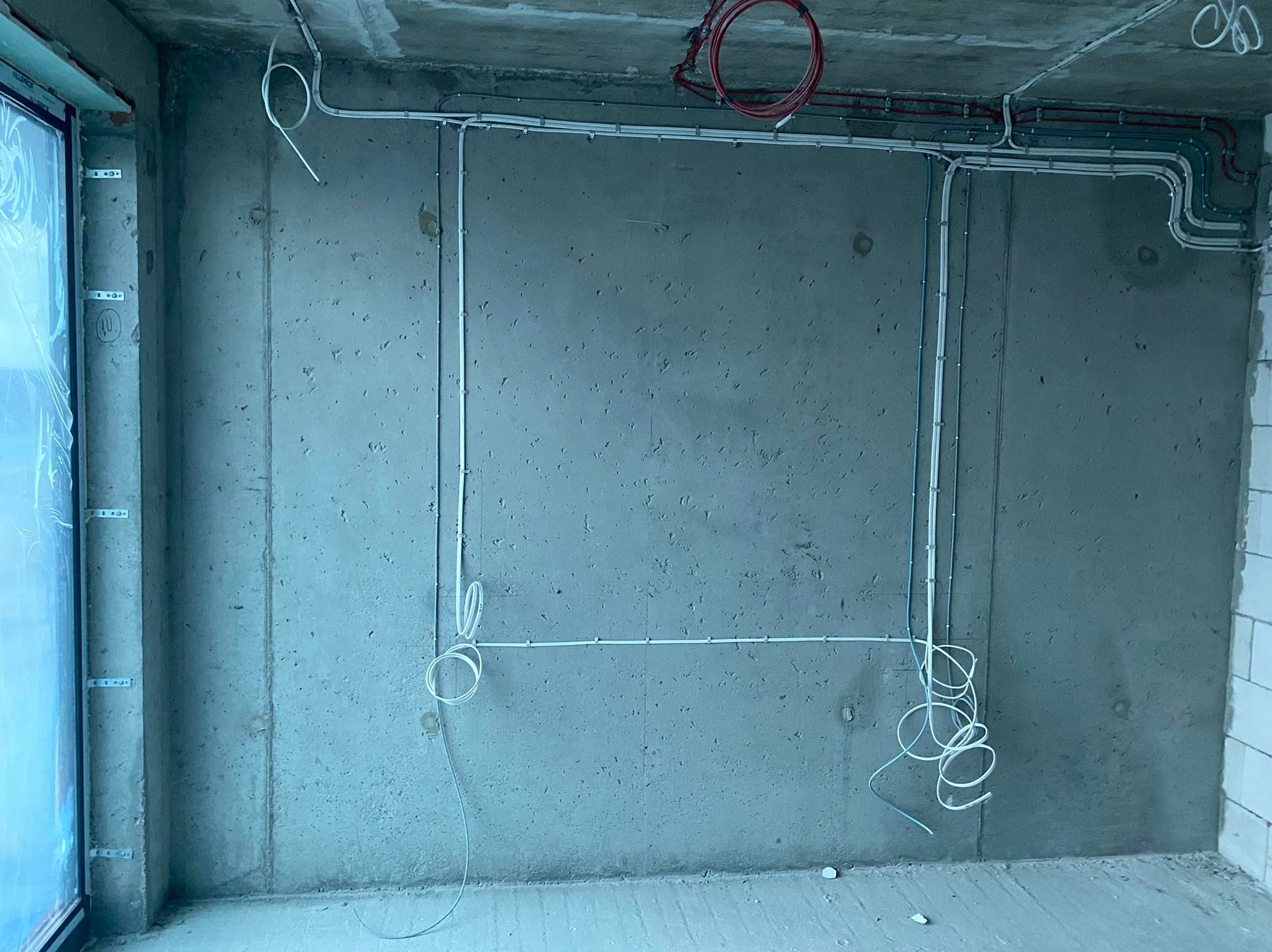 Surowe wnętrze w trakcie prac wykończeniowych z widocznymi przewodami elektrycznymi poprowadzonymi po ścianach i suficie, częściowo zamocowanymi, z widocznymi zwisającymi końcówkami kabli.