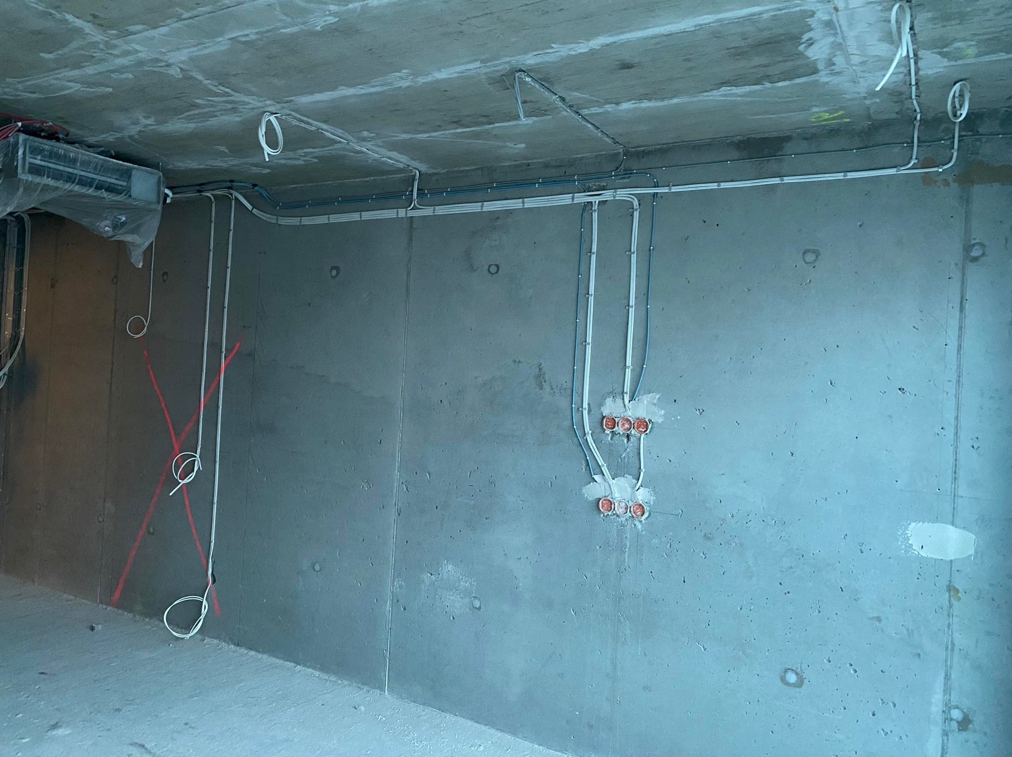 Surowy betonowy pokój z poprowadzonymi przewodami elektrycznymi w peszlach, puszkami pod gniazdka i wyłączniki oraz instalacją wentylacyjną pod sufitem w folii ochronnej.