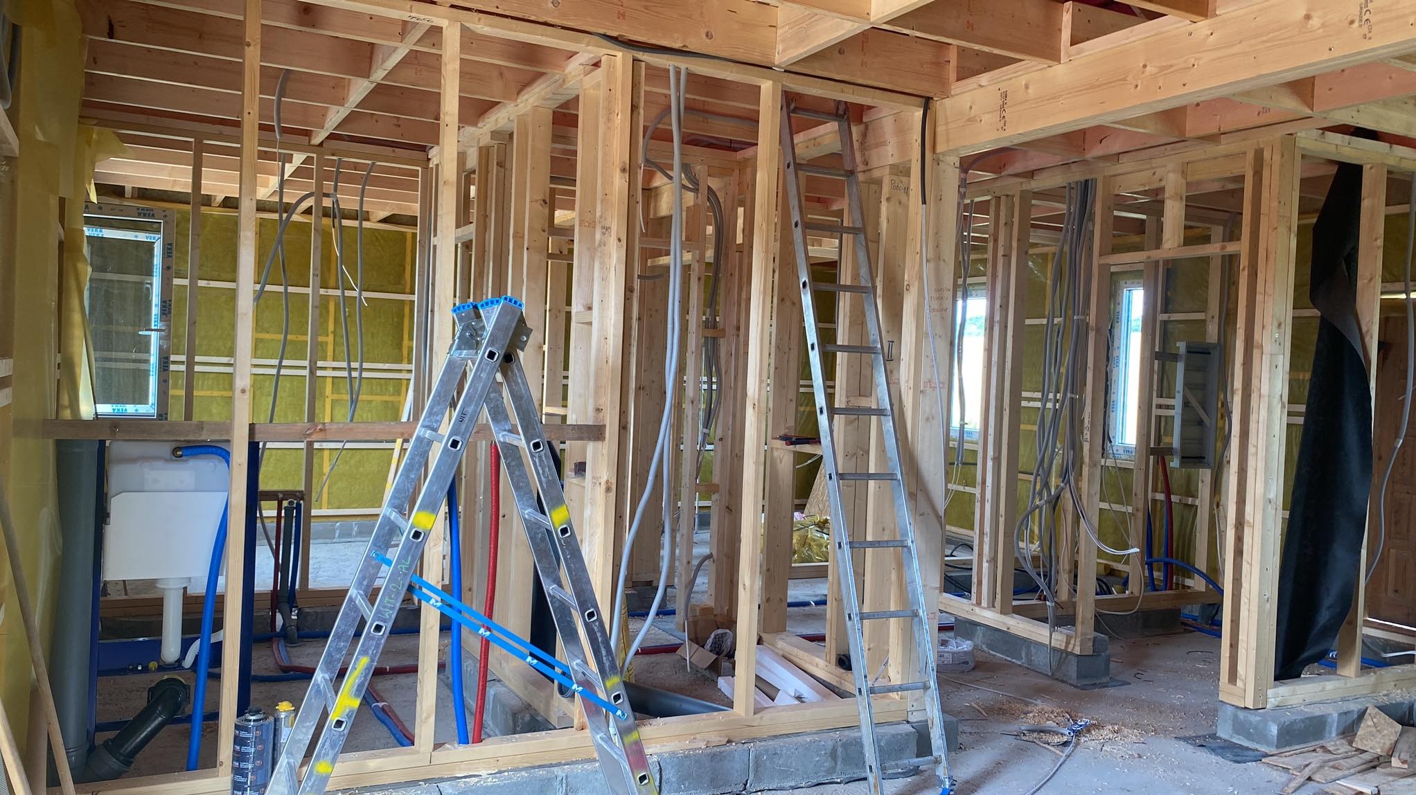 Wnętrze domu w trakcie budowy z widoczną drewnianą konstrukcją ścian, instalacją elektryczną w postaci szarych przewodów oraz rurami wodnymi w kolorze niebieskim i czerwonym, dwie aluminiowe...