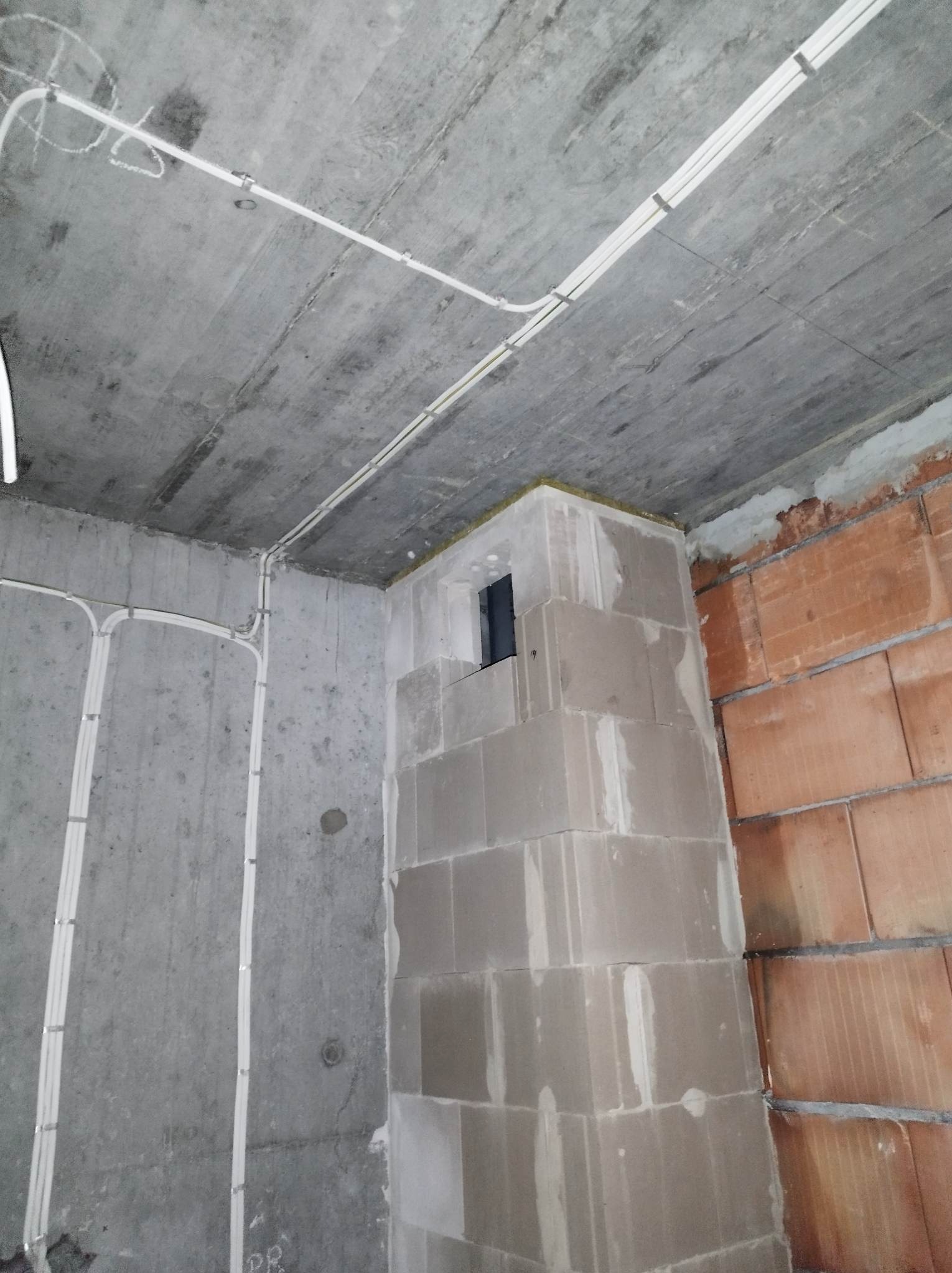 Surowy betonowy sufit z zamocowanymi białymi kablami elektrycznymi, ściany z betonu i czerwonej cegły w stanie surowym, widoczny komin z pustaków, instalacja w trakcie realizacji.