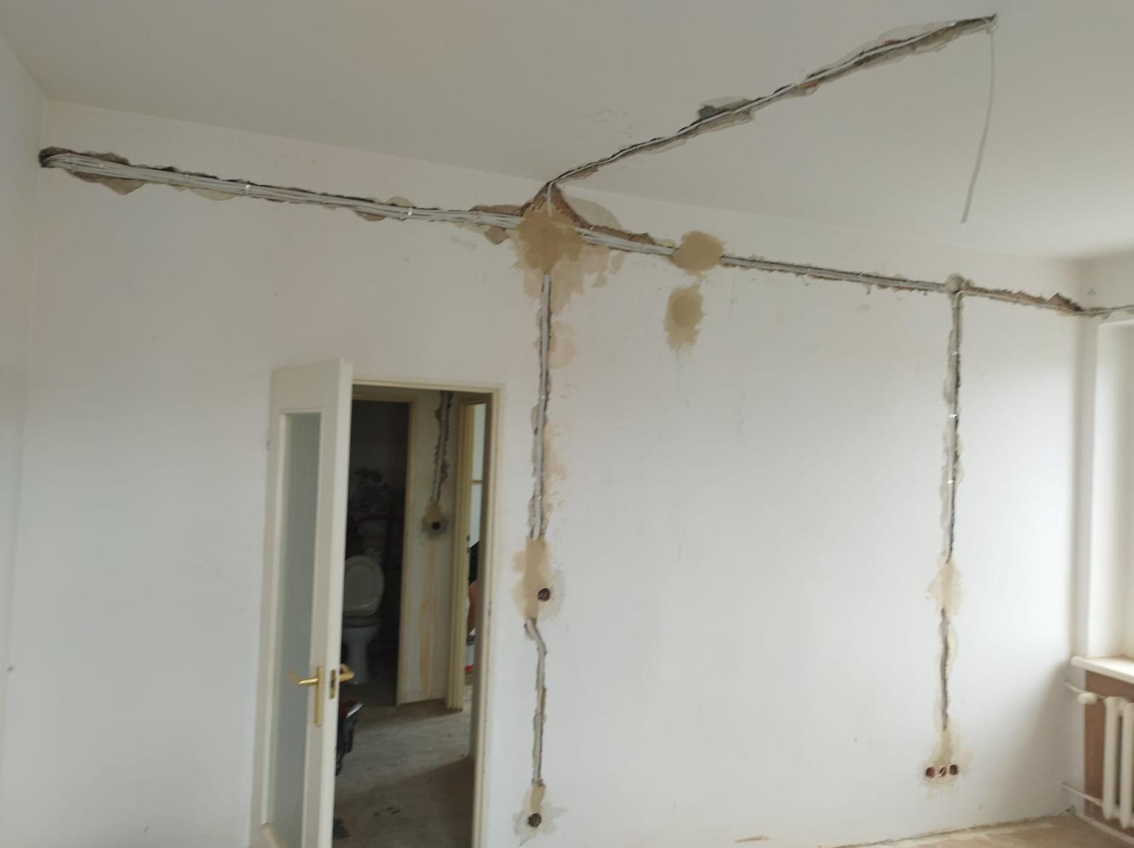 Przygotowanie do rozprowadzenia przewodów elektrycznych w nowym mieszkaniu: bruzdy w ścianach i suficie, widoczne przewody w peszlach, otwarte otwory pod gniazdka, surowy stan wykończenia wnętrza...