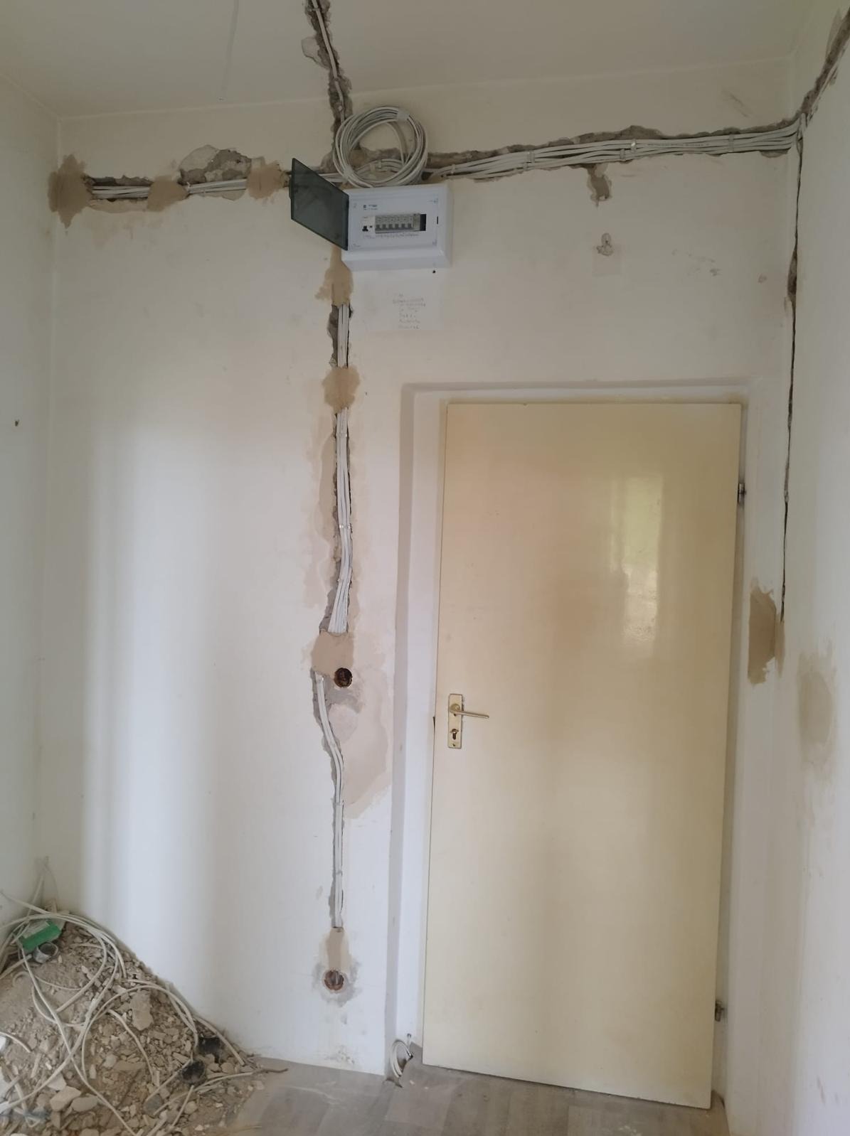 Wnętrze w trakcie remontu z widoczną, otwartą skrzynką bezpiecznikową, nowymi przewodami elektrycznymi w bruzdach na ścianie oraz stertą starych kabli na podłodze.