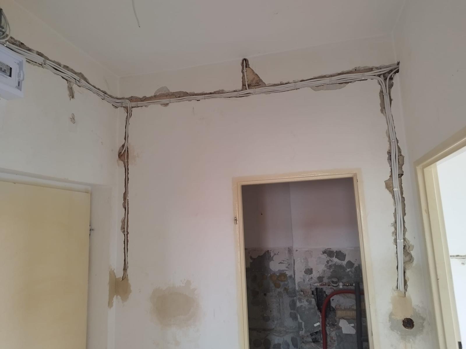 Wnętrze w trakcie remontu z widocznymi przewodami elektrycznymi ułożonymi w bruzdach na ścianach i pod sufitem, przygotowanymi do zamaskowania gipsem, widoczna skrzynka rozdzielcza i fragment...