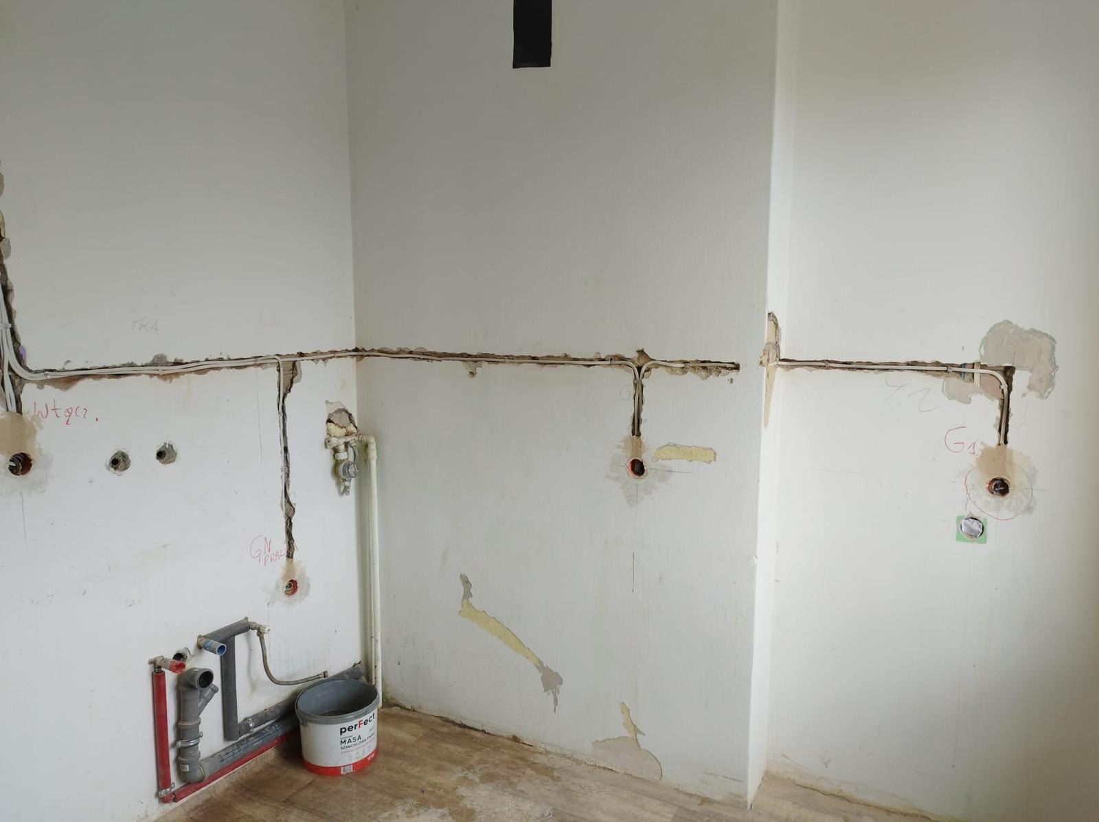Ściana w trakcie remontu z widocznymi przewodami elektrycznymi w bruzdach, instalacją wodną i wiaderkiem masy szpachlowej Perfect Haus. Na ścianie widoczne odręczne notatki.
