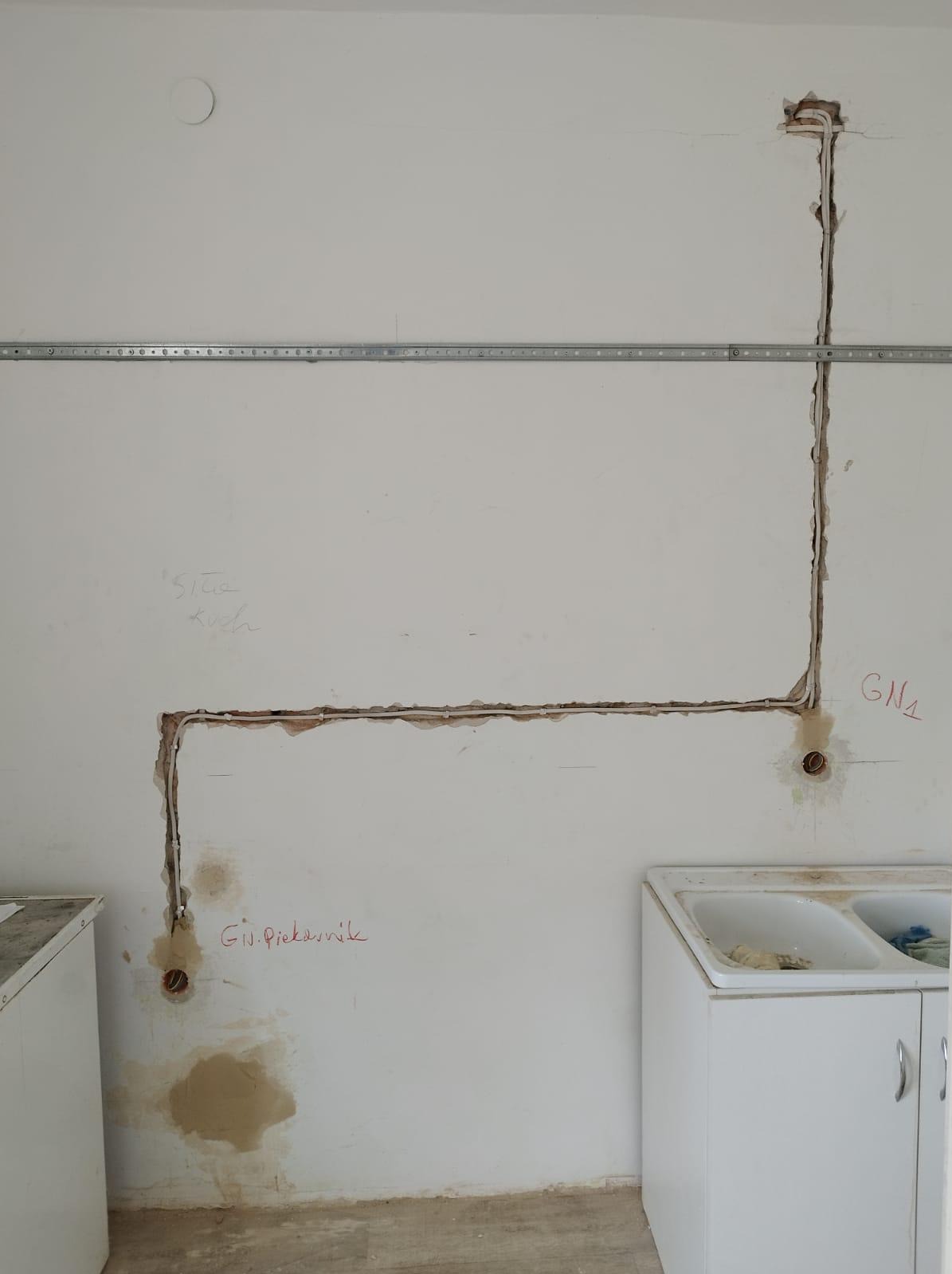 Biała ściana w trakcie remontu kuchni z wyżłobieniami pod przewody elektryczne, widoczne puszki instalacyjne, metalowa listwa montażowa i odręczne notatki na ścianie.