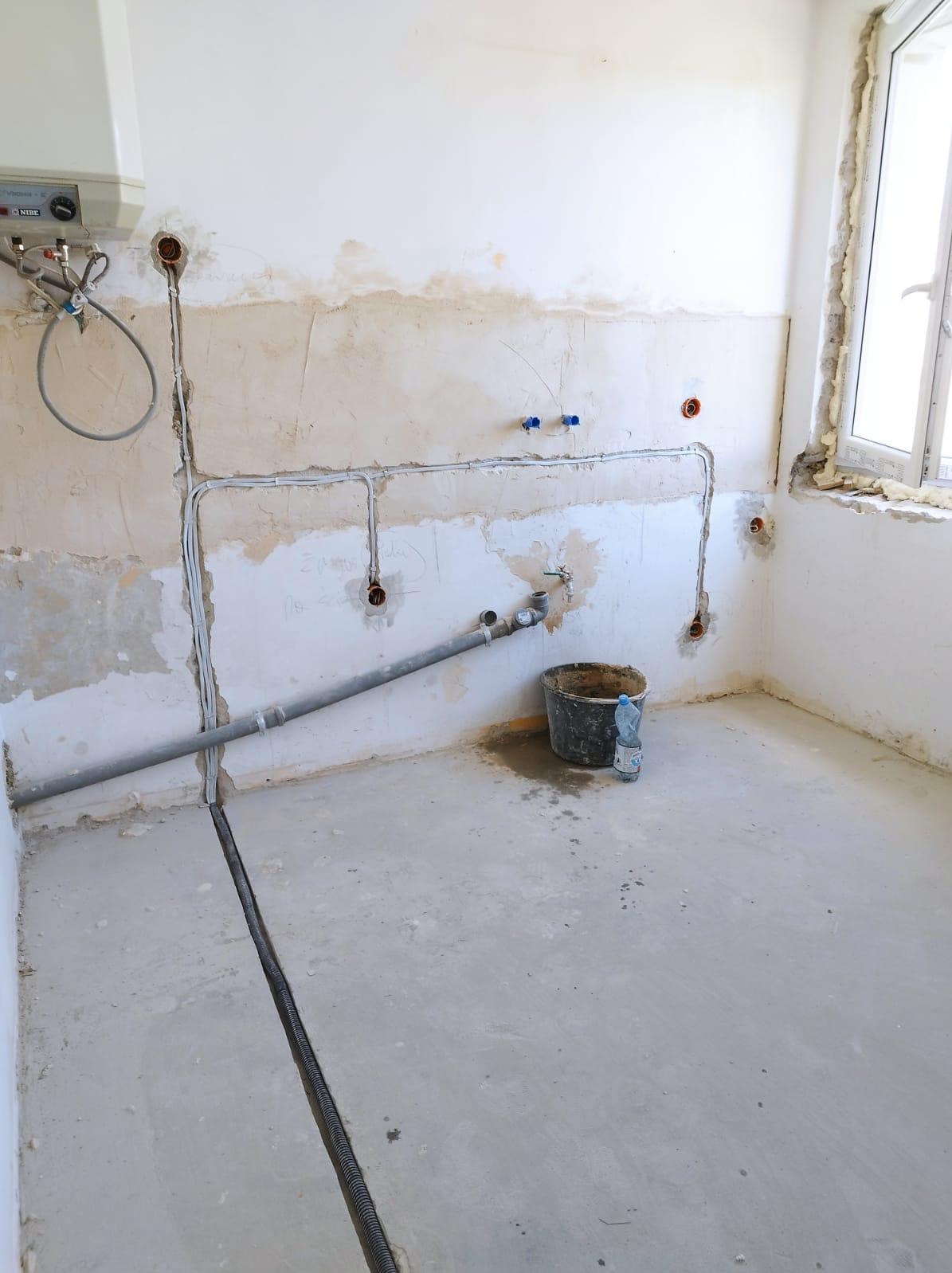 Surowe wnętrze w trakcie remontu: widoczne przewody elektryczne w ścianie, rury hydrauliczne, bojler, okno z pianką montażową i wiadro z wodą na betonowej podłodze.