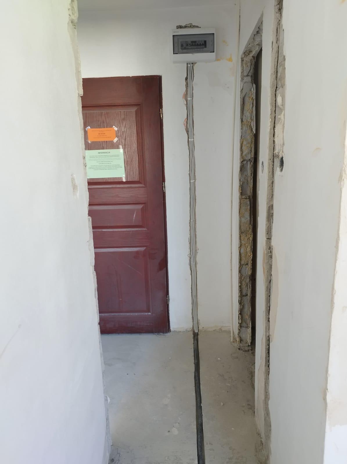 Rozdzielnica elektryczna natynkowa z przewodami w bruzdach ściennych, brązowe drzwi z informacją, surowy stan wykończenia wnętrza.