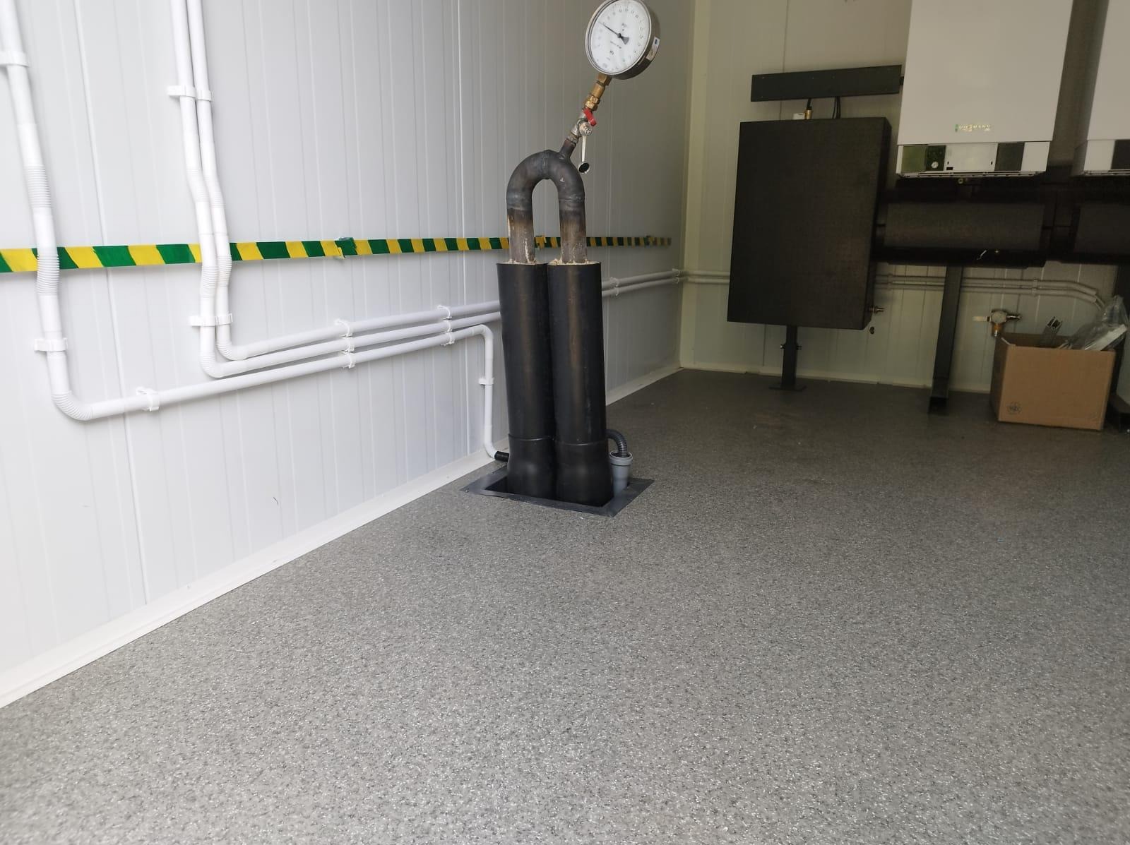 W kotłowni w Dzierzgoniu widoczny fragment instalacji z dwoma czarnymi rurami połączonymi u góry kolankiem z manometrem, na ścianie białe rury i żółto-zielony pas ostrzegawczy, w tle piec.