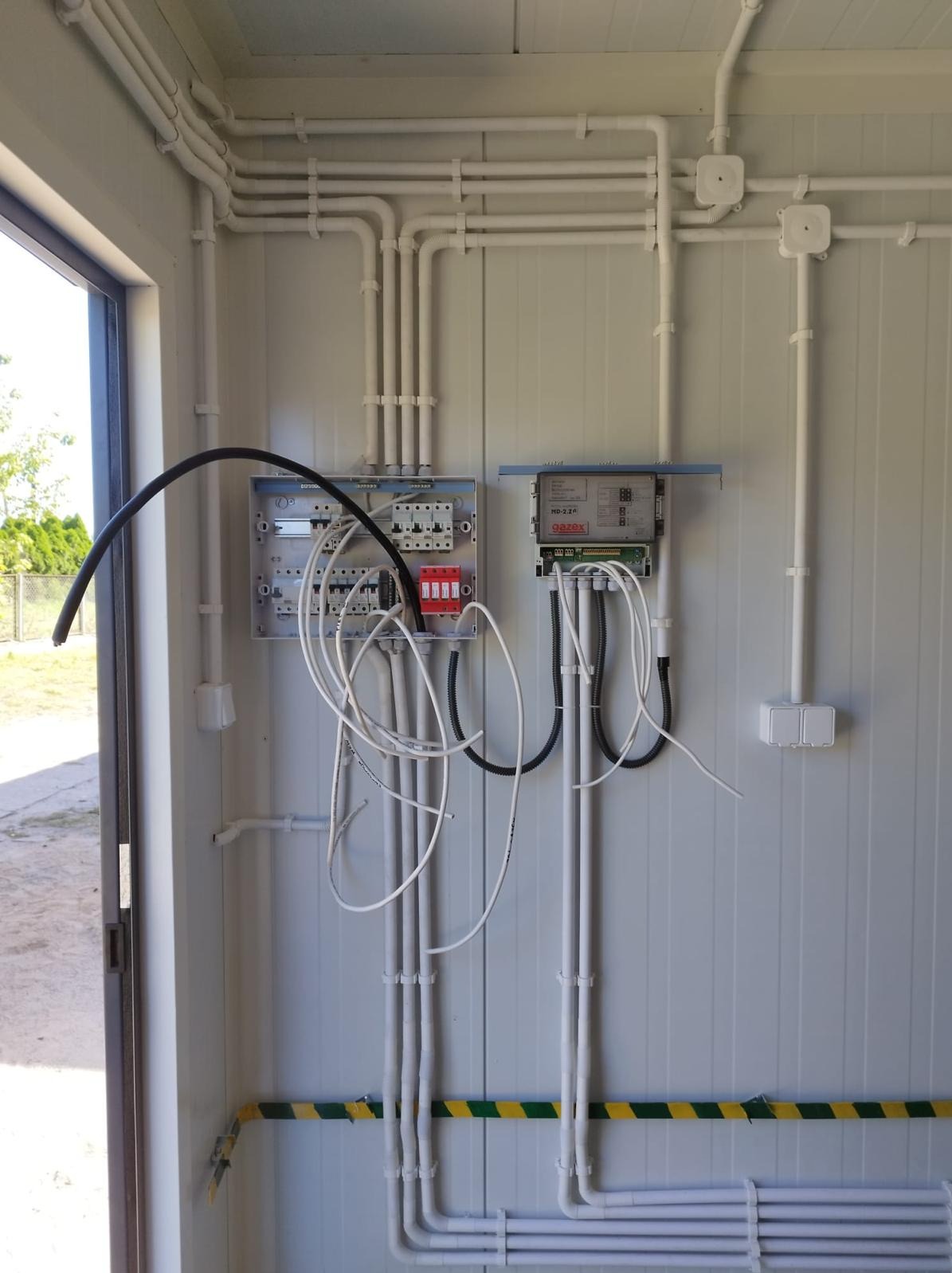 Wnętrze pomieszczenia z widoczną rozdzielnicą elektryczną, przewodami w białych rurkach PCV i czarnymi kablami, z żółto-czarną taśmą ostrzegawczą na dole ściany.