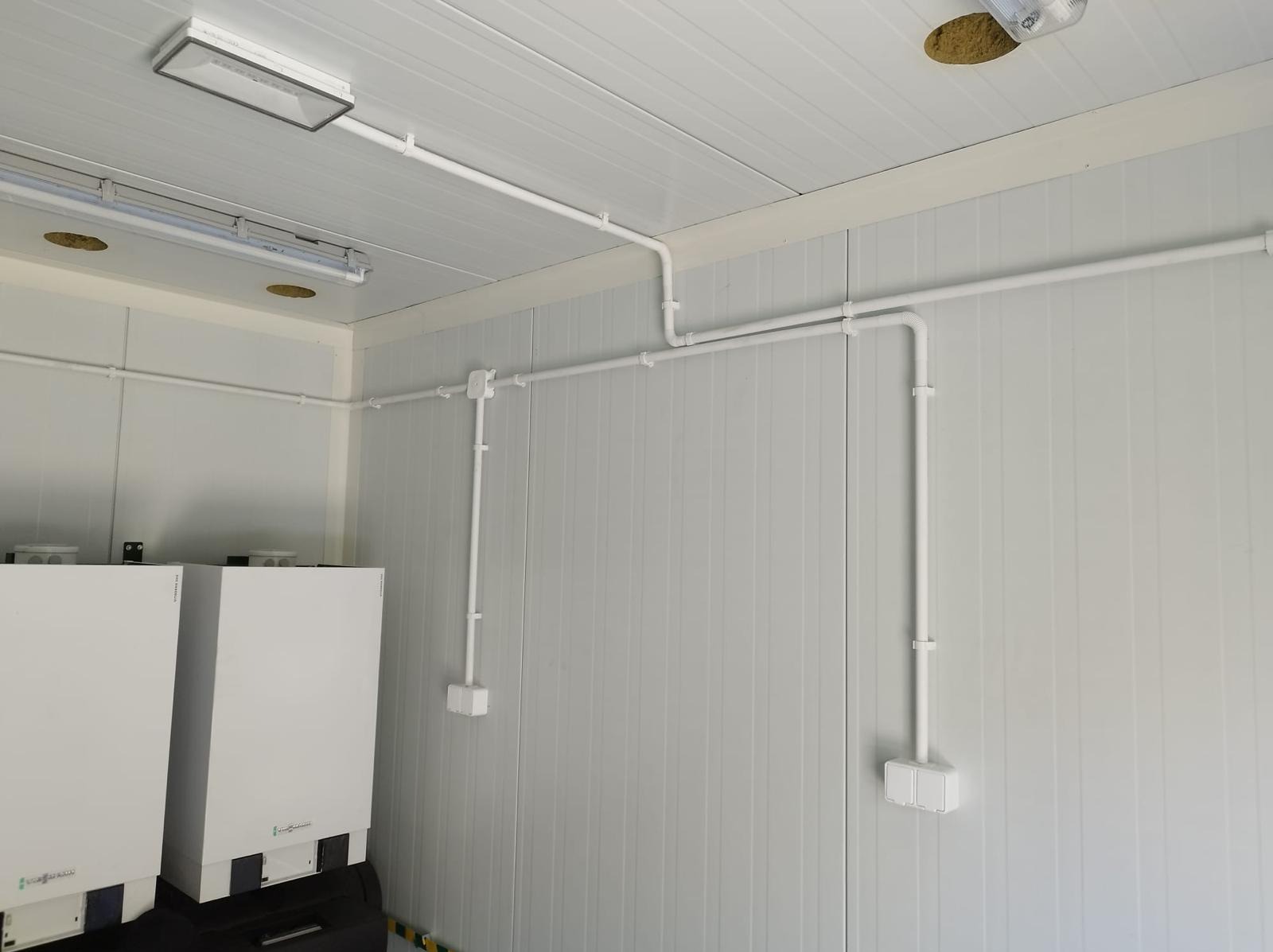 Białe rury i puszki instalacyjne na ścianie z paneli w chłodni, widoczne dwa białe urządzenia grzewcze Viessmann.
