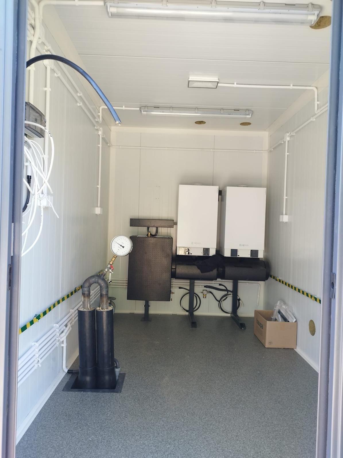 Wnętrze pomieszczenia technicznego z dwoma piecami grzewczymi, manometrem, instalacją rurową i elektryczną oraz kartonem Haupa na podłodze.