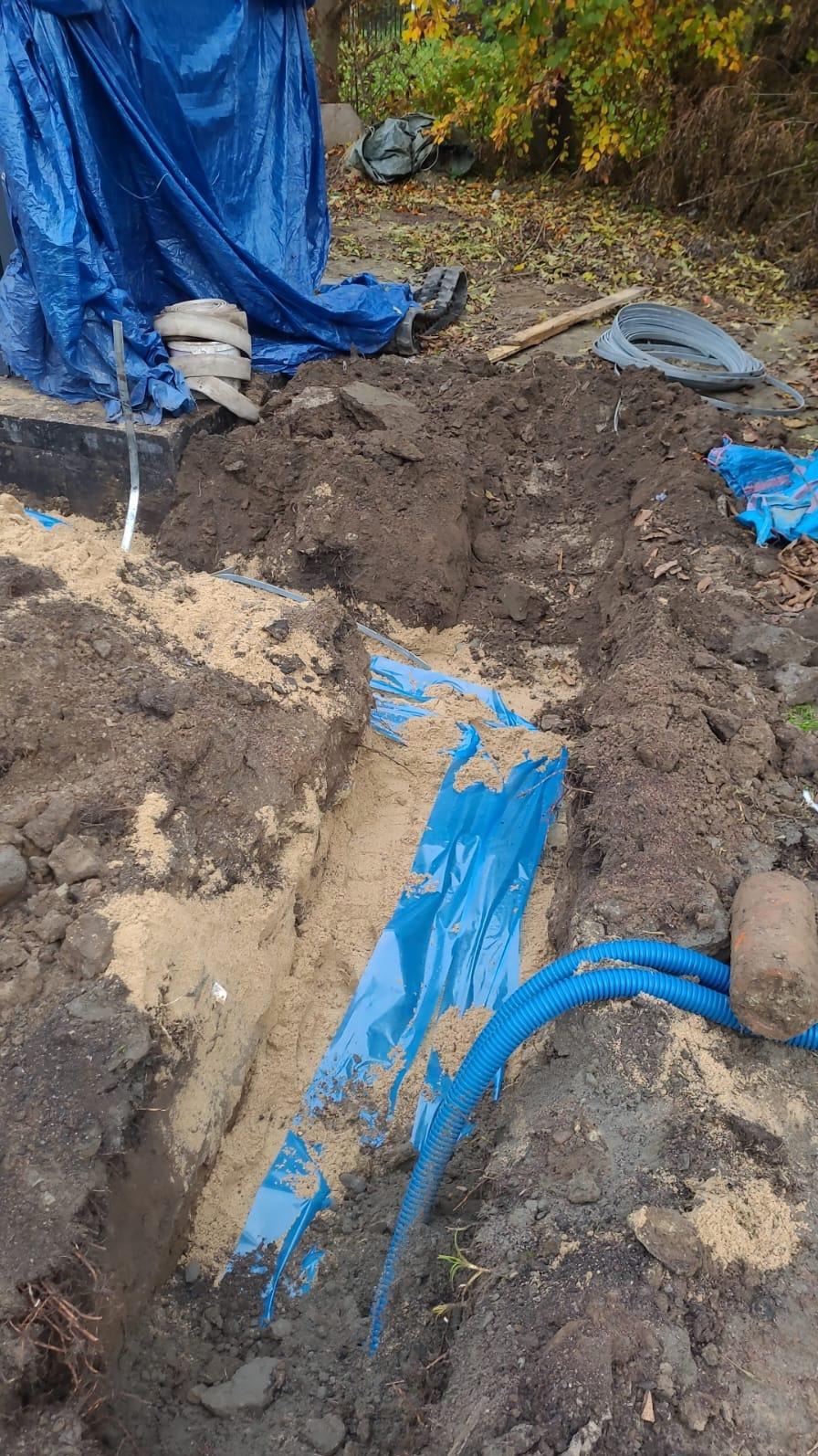 Wykop w ziemi wyłożony niebieską folią, widoczne niebieskie rury karbowane i przewody elektryczne, przygotowane do zakopania.