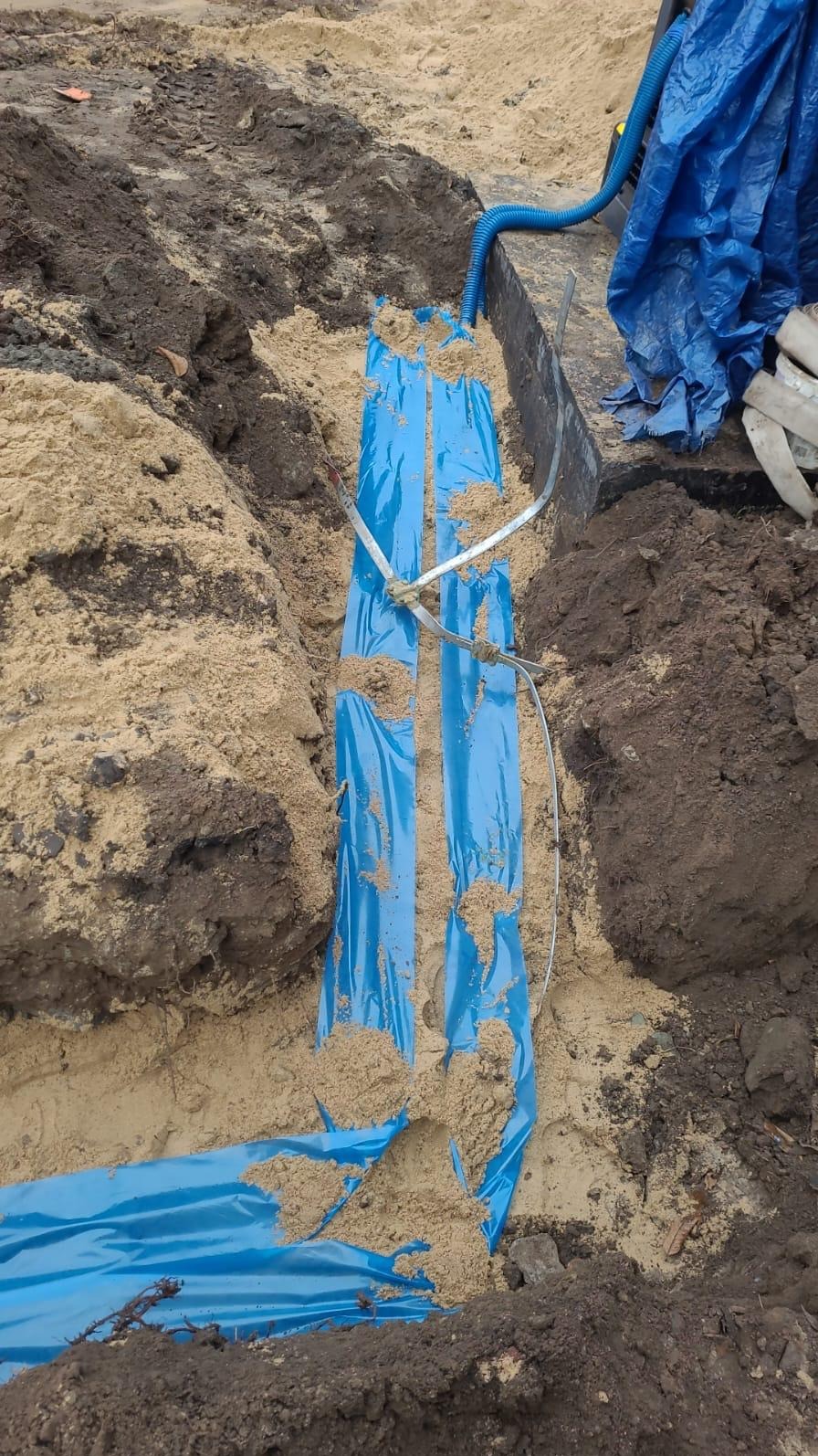 Wykop z piaskiem, w którym ułożono niebieską folię zabezpieczającą kable elektryczne, widoczne przewody i rura wentylacyjna.