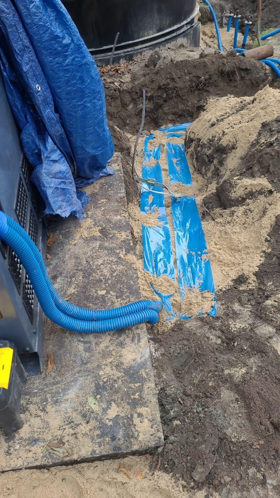 Wykop z piaskiem, w którym ułożono niebieskie rury osłonowe przewodów elektrycznych, widoczne kable, obok czarna skrzynka z żółtą naklejką i niebieski karbowany przewód.
