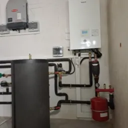 Nowoczesna instalacja grzewcza z białym piecem gazowym Panasonic, szarym zasobnikiem ciepłej wody i czerwonym naczyniem przeponowym, zamontowana na białej ścianie w kotłowni.