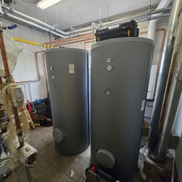 INSTALACJE KOMPLEKS SPÓŁKA Z OGRANICZONĄ ODPOWIEDZIALNOŚCIĄ - Dwa szare zbiorniki buforowe Galmet w kotłowni, widoczne rury miedziane, skrzynka narzędziowa na jednym ze zbiorników, instalator w trakcie pracy.