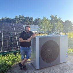 INSTALACJE KOMPLEKS SPÓŁKA Z OGRANICZONĄ ODPOWIEDZIALNOŚCIĄ - Uśmiechnięty mężczyzna opiera się o nowoczesną pompę ciepła na zewnątrz, z panelami słonecznymi w tle i błękitnym niebem. Widoczne oznaczenia efektywności energetycznej.