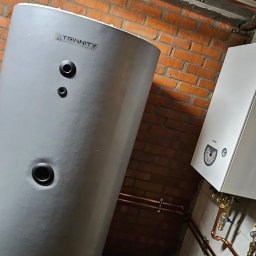 INSTALACJE KOMPLEKS SPÓŁKA Z OGRANICZONĄ ODPOWIEDZIALNOŚCIĄ - Szary zasobnik ciepłej wody użytkowej Trinity obok białego pieca gazowego Fermet, z miedzianymi rurami i ceglaną ścianą w tle. Widoczny łuk sportowy na ścianie.