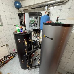 INSTALACJE KOMPLEKS SPÓŁKA Z OGRANICZONĄ ODPOWIEDZIALNOŚCIĄ - Instalacja kotłowni z dwoma zbiornikami, czarnym i srebrnym Viqtis, oraz widoczną instalacją hydrauliczną i narzędziami na podłodze w pomieszczeniu wyłożonym kafelkami.