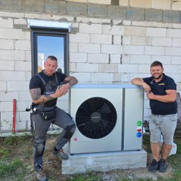 INSTALACJE KOMPLEKS SPÓŁKA Z OGRANICZONĄ ODPOWIEDZIALNOŚCIĄ - Dwóch instalatorów pozuje obok zamontowanej pompy ciepła Kolton na betonowym fundamencie, na tle ściany budynku z białego bloczka i okna z roletą.