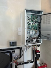 Otwarty panel serwisowy nowoczesnego pieca gazowego wiszącego na ścianie, widoczne przewody, elektronika i elementy instalacji grzewczej, z podłączonymi rurami miedzianymi i elementami hydraulicznymi.