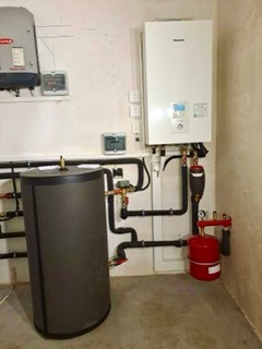 Kotłownia z zasobnikiem ciepłej wody, piecem gazowym i rozbudowaną instalacją hydrauliczną z czarnych rur. Widoczny regulator temperatury i panel solarny na ścianie.