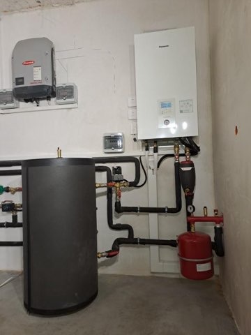 Nowoczesna instalacja grzewcza z białym piecem gazowym Panasonic, szarym zasobnikiem ciepłej wody i czerwonym naczyniem przeponowym, zamontowana na białej ścianie w kotłowni.