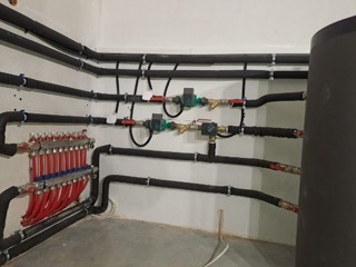 Rozdzielacz ogrzewania podłogowego z czerwonymi rurkami, widoczne zawory i izolowane rury doprowadzające, zamocowane do ściany w surowym, jasnym pomieszczeniu.