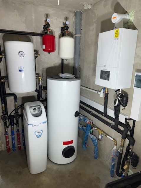 Kompletna kotłownia w surowym pomieszczeniu: biały piec gazowy, zbiorniki, filtry i rury. Widoczne czarne rury instalacyjne.