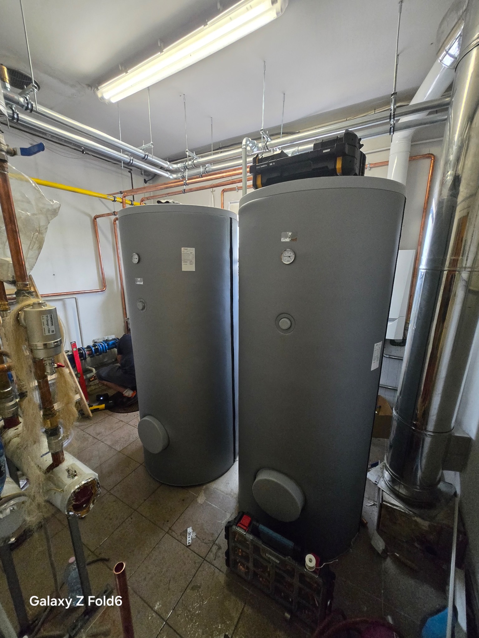 Dwa szare zbiorniki buforowe Galmet w kotłowni, widoczne rury miedziane, skrzynka narzędziowa na jednym ze zbiorników, instalator w trakcie pracy.