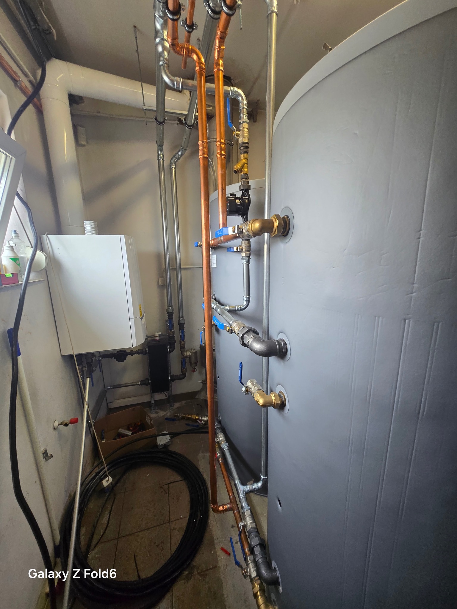 Instalacja CO z piecem gazowym i zasobnikiem CWU. Miedziane rury, zawory, biały piec wiszący. Widok na kotłownię z elementami instalacji grzewczej.