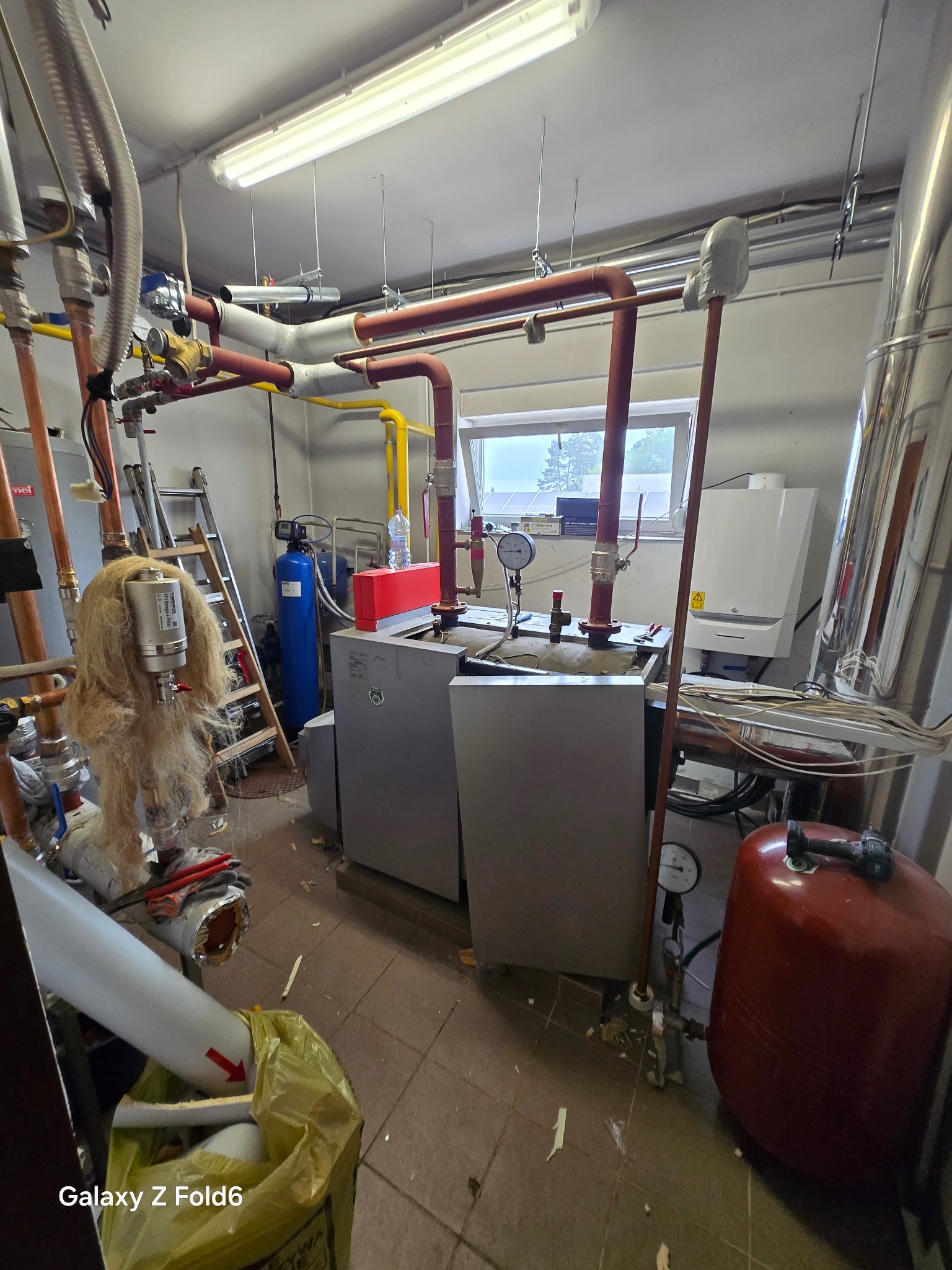 Kotłownia z dwoma piecami, rurami miedzianymi i żółtymi, zbiornikiem wyrównawczym, filtrem wody i drabiną. Widoczne okno i narzędzia.