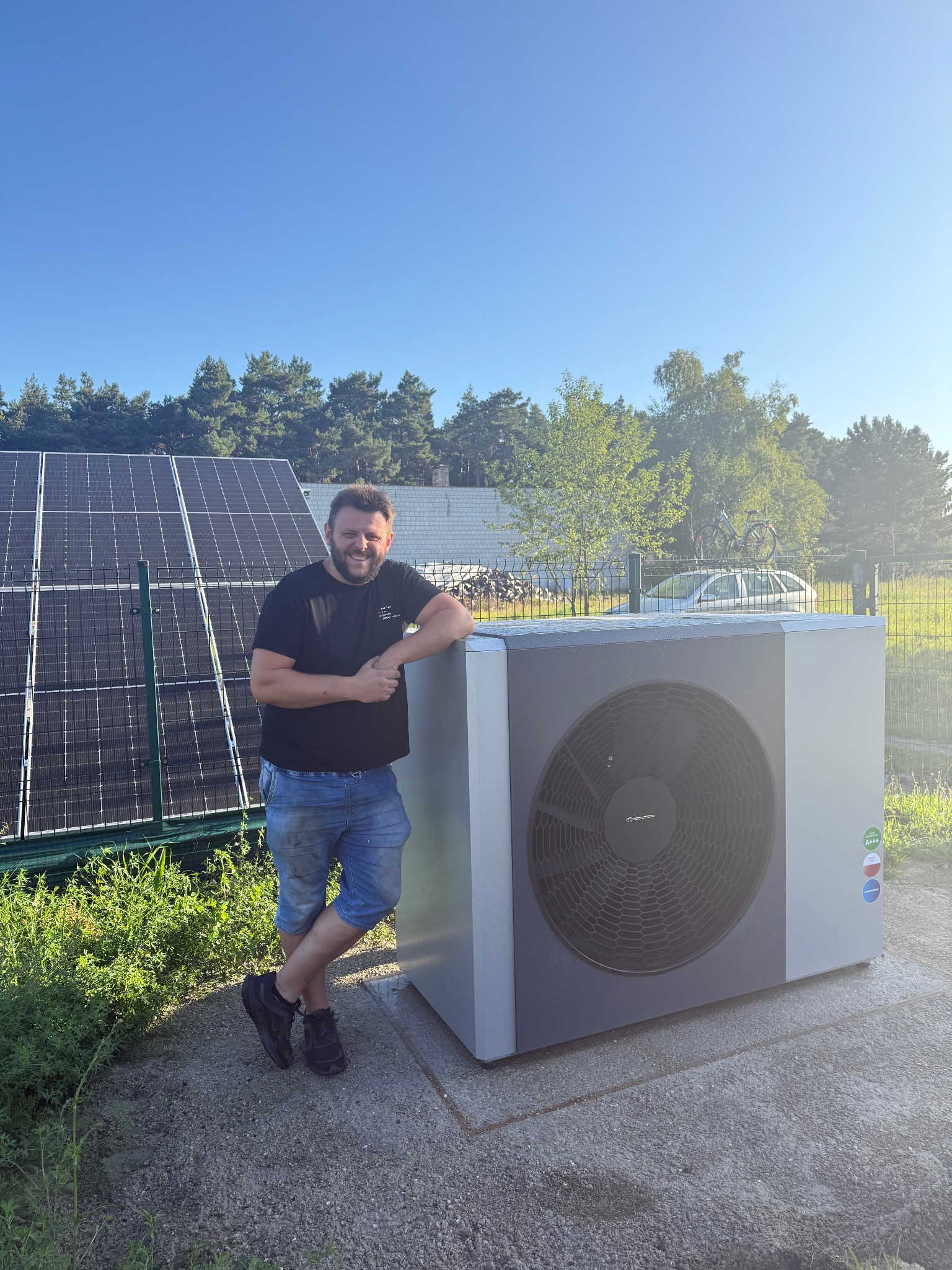 Uśmiechnięty mężczyzna opiera się o nowoczesną pompę ciepła na zewnątrz, z panelami słonecznymi w tle i błękitnym niebem. Widoczne oznaczenia efektywności energetycznej.