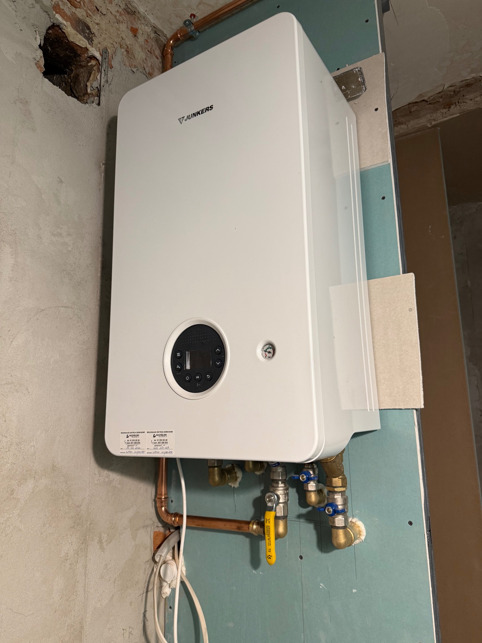 Biały piec gazowy Junkers zamontowany na ścianie z widocznymi rurami miedzianymi i zaworami. Ściana częściowo wykończona płytą gipsową, widoczne ślady po instalacji.