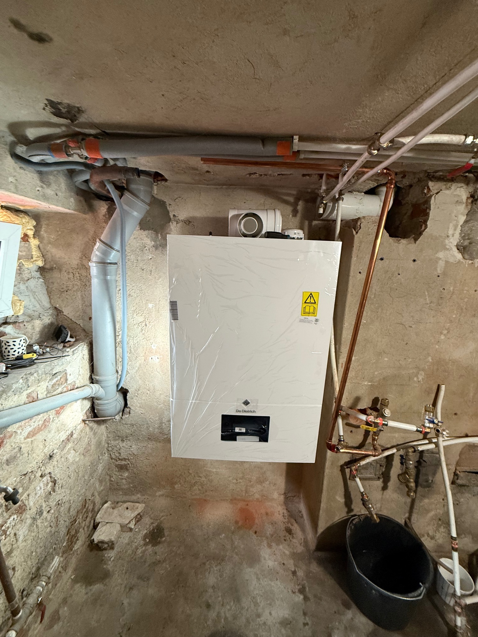 Nowy, biały piec gazowy marki De Dietrich, jeszcze w folii ochronnej, zamontowany w surowym pomieszczeniu z widocznymi rurami i instalacją hydrauliczną.