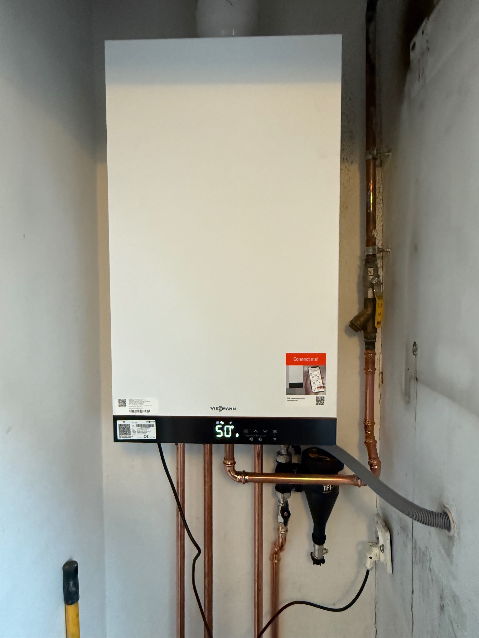 Biały, wiszący piec gazowy Viessmann z miedzianą instalacją, zamontowany w pomieszczeniu z szarymi ścianami. Widoczny filtr TF1 i fragment narzędzia w lewym dolnym rogu.