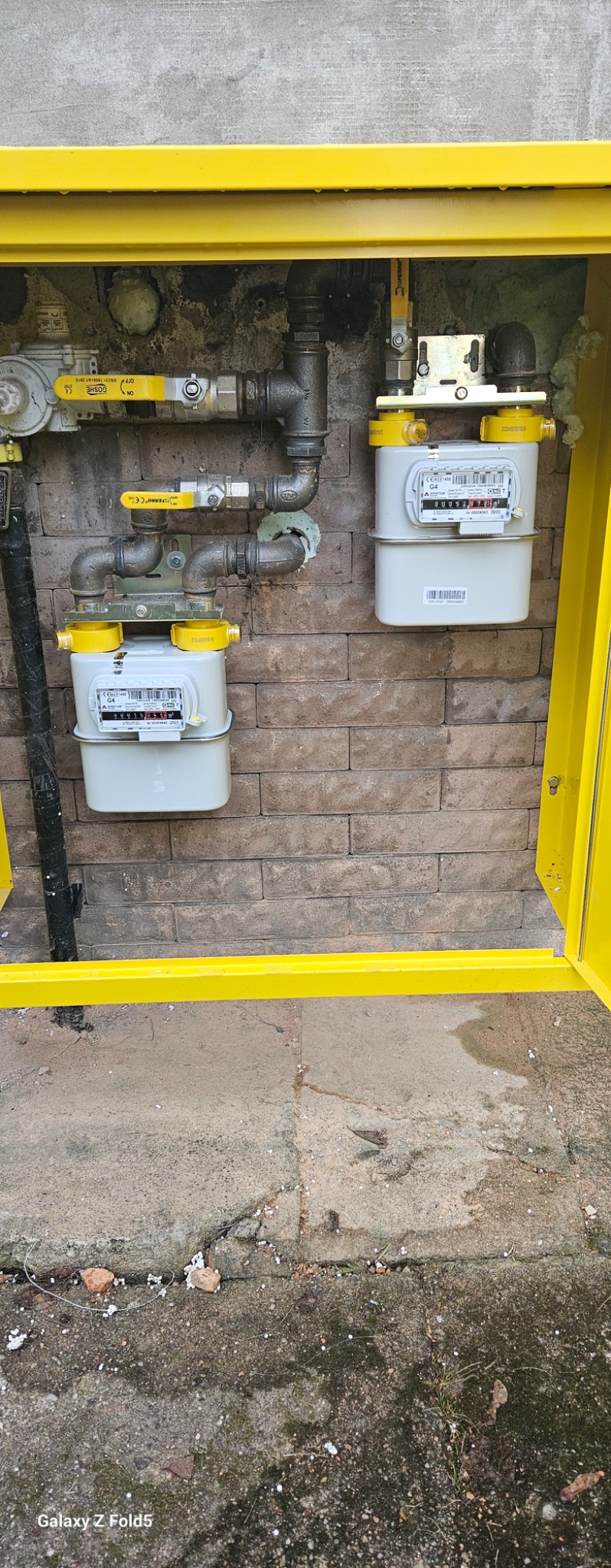 Dwa gazomierze w żółtej szafce na ceglanej ścianie. Widoczne rury i zawory. Instalacja gazowa po montażu, z bliska.