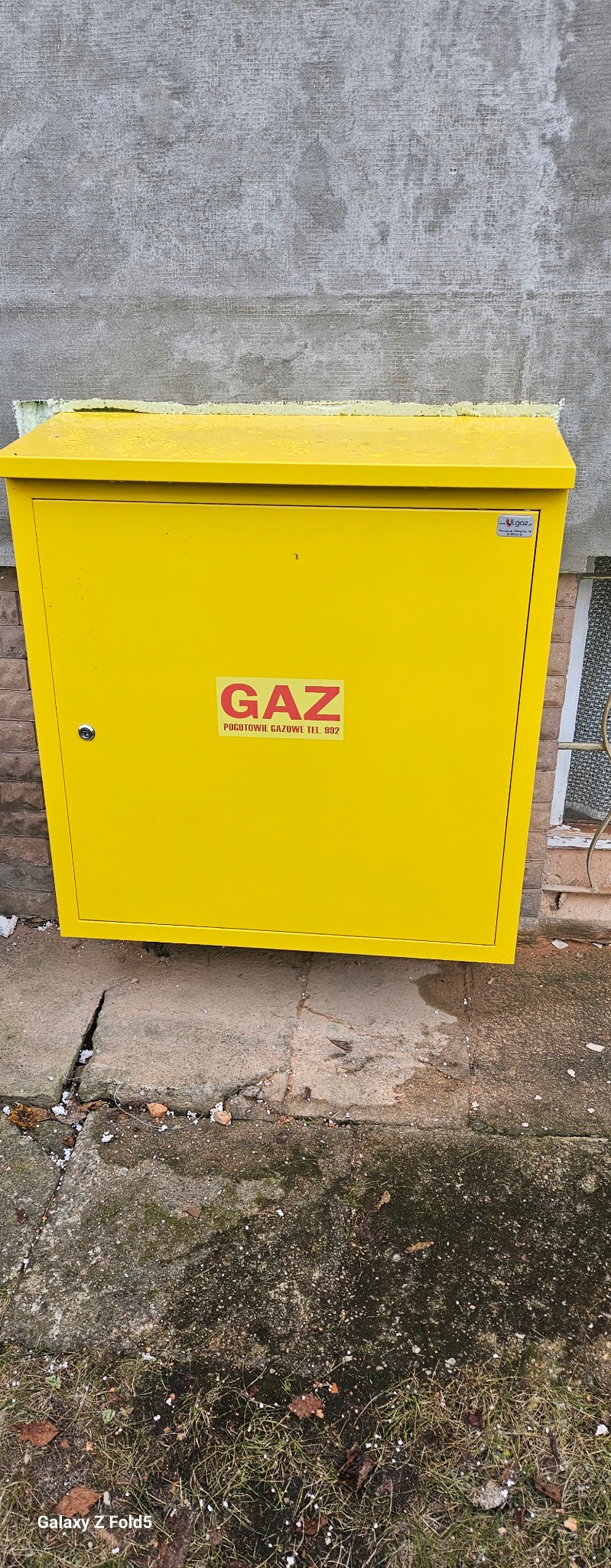 Żółta skrzynka gazowa na zewnątrz budynku, z naklejką 'GAZ Pogotowie Gazowe'. Widoczne ślady użytkowania i otoczenie: betonowa podstawa, fragment ściany i okna.